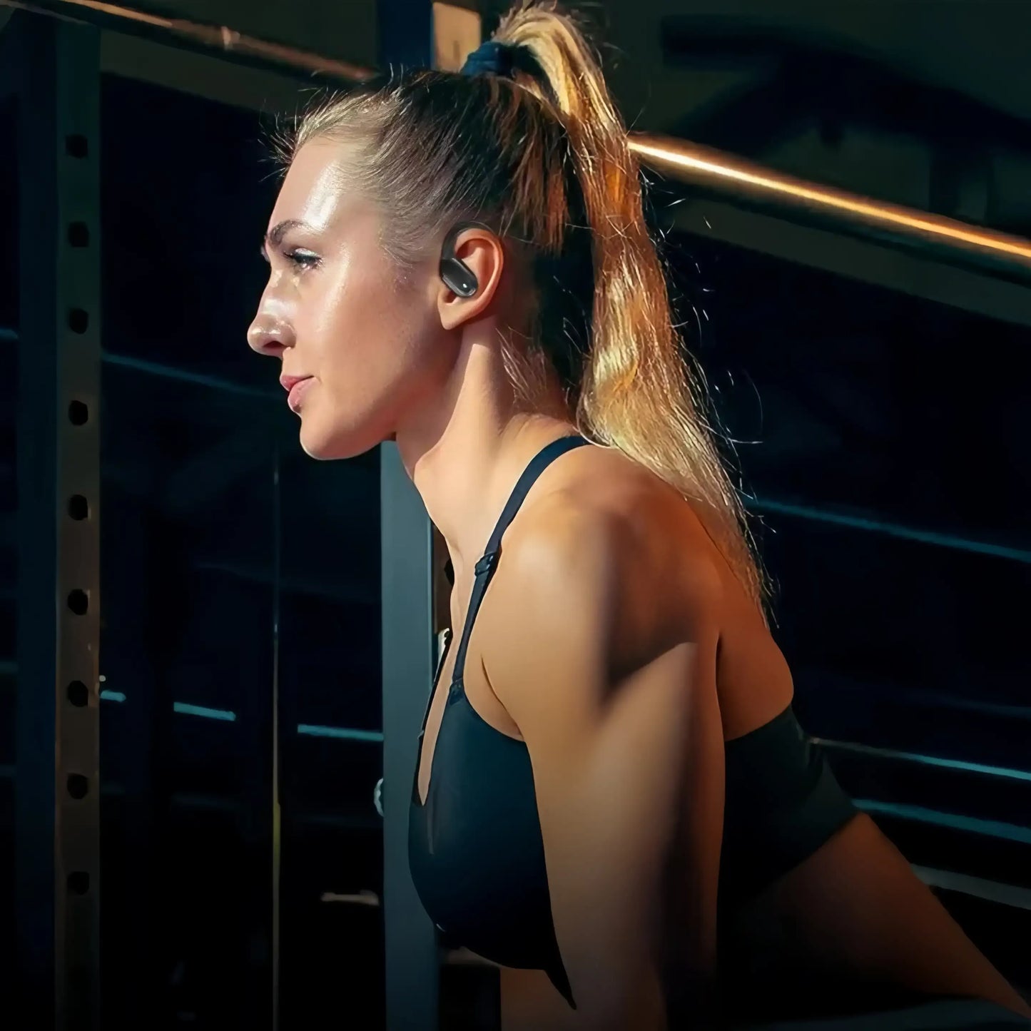 A woman in a gym wearing a black sports bra and a black Tekarvo Wireless Sports Earbuds, shown in profile against a dark gym background with a power rack.